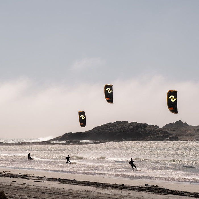 Three kite surfers with Code NXT v2 kites in the sky over a beach and ocean landscape.