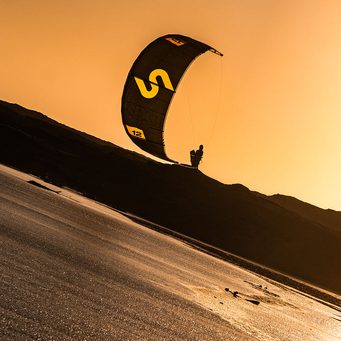 Person kiteboarding on a beach with a large Code NXT v2 kite against an orange sky.